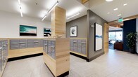 This thoughtfully designed mail room offers a clean, contemporary aesthetic that balances functionality with visual appeal. The space is anchored by custom-built light wood cabinetry and countertops that house an extensive array of individual resident mailboxes and parcel lockers, all finished in a coordinated brushed gray tone. The warm, natural wood tones of the cabinetry contrast elegantly with the muted gray accent walls, creating a refined and inviting atmosphere.

Linear pendant lighting runs along the white ceiling, providing bright, even illumination throughout the room and enhancing the sense of openness. The light-toned large-format tile flooring adds to the airy quality of the space, reflecting light and maintaining a cohesive, modern palette.

Framed artwork is thoughtfully placed throughout the room, featuring abstract and landscape-inspired pieces that elevate the environment beyond a purely utilitarian function. A decorative sculptural accent piece rests atop one of the central countertops, adding a curated, design-forward touch.

The layout is organized with central island-style mailbox stations complemented by wall-mounted units, allowing for efficient circulation and easy access. Overall, this mail room exemplifies a high standard of communal amenity design, combining practicality with a polished, welcoming ambiance.
