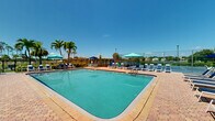 Located at 222 Kingfisher Way in Royal Palm Beach, Florida, this outdoor community amenity space is designed to deliver a resort-style experience in a sunny South Florida setting. At its center is a generously sized swimming pool with crystal-clear turquoise water, surrounded by a wide paver deck that accommodates numerous lounge chairs and shade umbrellas, ideal for relaxing under the abundant Florida sunshine. A separate spa is tucked into one corner of the deck, offering a more intimate retreat alongside the main pool. Adjacent to the pool area, a dedicated outdoor lounge and BBQ station provides tables, chairs, and grilling facilities, perfect for casual gatherings and outdoor dining framed by tall swaying palm trees. A children's playground is situated nearby, making the space well-suited for families. The pool area is enclosed by a wrought iron fence that maintains safety while preserving unobstructed views of the serene lake beyond, creating a tranquil waterfront backdrop. A pool house structure on the property provides convenient access to facilities. Lush tropical landscaping, including mature palms and manicured greenery, enhances the overall ambiance, giving the space a vibrant, resort-like atmosphere throughout.