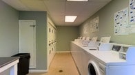 This shared laundry facility is designed for practical, efficient use. The room is finished with soft sage-green walls and neutral carpeted flooring, creating a clean and functional environment. Overhead, recessed panel lighting provides bright, even illumination throughout the space. Along one wall, a row of top-loading washing machines is neatly arranged, offering ample capacity for multiple users simultaneously. The opposite wall features stacked dryer units, maximizing the use of vertical space and increasing the overall number of available machines. Instructional signage is posted on the walls, offering guidance on machine usage and laundry best practices. A white-painted door provides access to an adjoining area, adding to the room's accessibility. The layout is straightforward and organized, allowing users to move comfortably between machines. Overall, the facility is well-maintained and thoughtfully arranged to serve the laundering needs of a multi-unit residential building efficiently.