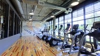 This fitness center showcases a compelling blend of industrial and contemporary design elements. The exposed concrete ceiling with visible ductwork and conduit gives the space an urban, warehouse-inspired aesthetic, complemented by pendant-style industrial lighting that provides warm, functional illumination throughout.

A defining feature of the space is the expansive floor-to-ceiling window wall along one side, flooding the interior with abundant natural light and offering open views of the surrounding greenery. This connection to the outdoors enhances the energy of the workout environment.

The equipment layout is well-organized, with a row of treadmills and elliptical machines positioned to face the windows, allowing users to enjoy the exterior views during cardio sessions. Each machine is equipped with an individual monitor for entertainment or workout tracking.

The hardwood flooring features a distinctive multi-colored pattern with blue, red, and natural wood tones, adding a dynamic visual element underfoot. Anchoring the opposite wall is a large-scale artistic mural with an abstract, nature-inspired design, providing a creative focal point that enlivens the space and reinforces its modern character.