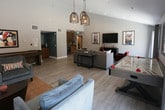 This inviting single-floor community lounge is designed for both relaxation and entertainment, offering a thoughtfully curated blend of modern and rustic elements throughout. The main living area is generously proportioned, featuring light wood-look flooring, vaulted ceilings, and recessed lighting complemented by striking woven pendant light fixtures that add warmth and character. Large sliding glass doors flood the space with natural light and provide views of the outdoor surroundings, creating an airy, open atmosphere. Comfortable sectional and accent seating in neutral tones with bold orange and red accents invite gathering, while a wall-mounted flat-screen TV anchors the lounge area. The game zone is a standout feature, equipped with a full-size air hockey table, a shuffleboard table, and a large decorative Scrabble board mounted on the wall, offering ample entertainment options. A dedicated seating nook with plush armchairs provides a quieter retreat alongside the games. The wet bar area is a design highlight, showcasing warm wood cabinetry, sleek gray countertops, a utility sink with an oil-rubbed bronze faucet, a beverage refrigerator, and a dramatic exposed brick accent wall adorned with floating wood shelves. Retro-inspired artwork and décor throughout the space lend a playful, eclectic personality to this well-appointed communal gathering area.