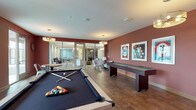 This single-floor game and entertainment room is designed with a vibrant, social atmosphere in mind. The space is defined by rich, deep red accent walls that create a warm, energetic ambiance throughout. Large-format tile flooring with a stone-like finish runs across the entire room, providing a durable yet visually appealing foundation.

At the center of the space sits a full-size pool table with a light wood frame and dark felt surface, positioned beneath a pair of contemporary multi-bulb pendant chandeliers that cast a warm, focused glow. Along one wall, a full-length shuffleboard table is accompanied by a collection of framed vintage-style artwork, reinforcing the playful character of the room. A wall-mounted flat-screen television flanked by sconce lighting offers an additional entertainment focal point.

The lounge area features a pair of upholstered accent chairs and a floor lamp, providing comfortable seating for those not actively playing. A small dining table with metal chairs sits near glass sliding doors that open to an outdoor balcony, allowing natural light to filter in. Additional glass panel partitions connect this space to an adjacent area, maintaining an open, connected feel while defining distinct zones within the overall layout.