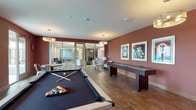 This single-floor game and entertainment room is designed with a vibrant, social atmosphere in mind. The space is defined by rich, deep red accent walls that create a warm, energetic ambiance throughout. Large-format tile flooring with a stone-like finish runs across the entire room, providing a durable yet visually appealing foundation.

At the center of the space sits a full-size pool table with a light wood frame and dark felt surface, positioned beneath a pair of contemporary multi-bulb pendant chandeliers that cast a warm, focused glow. Along one wall, a full-length shuffleboard table is accompanied by a collection of framed vintage-style artwork, reinforcing the playful character of the room. A wall-mounted flat-screen television flanked by sconce lighting offers an additional entertainment focal point.

The lounge area features a pair of upholstered accent chairs and a floor lamp, providing comfortable seating for those not actively playing. A small dining table with metal chairs sits near glass sliding doors that open to an outdoor balcony, allowing natural light to filter in. Additional glass panel partitions connect this space to an adjacent area, maintaining an open, connected feel while defining distinct zones within the overall layout.