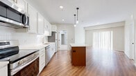 This thoughtfully designed 2-bedroom, 1-bathroom residence offers a functional and modern layout across a single floor. The open-concept kitchen and dining area serves as the heart of the home, featuring crisp white shaker cabinetry, a classic subway tile backsplash, quartz countertops, and a full suite of stainless steel appliances including a refrigerator, range, microwave, and dishwasher. A warm-toned wood center island with a marble-style countertop provides additional prep space and anchors the room beneath stylish pendant lighting. Recessed lighting and wood-look flooring extend throughout the main living areas, creating a bright and cohesive aesthetic. A large sliding glass door with vertical blinds allows natural light to fill the living area and offers access to the outdoors. The two bedrooms are finished with plush carpeting and feature large windows fitted with horizontal blinds. The primary bedroom includes a dedicated walk-in closet equipped with wire shelving and hanging rods for generous storage. The full bathroom is cleanly appointed with a white vanity, quartz countertop, and a tub-shower combination. Throughout the space, neutral tones and modern finishes create a clean, contemporary atmosphere.