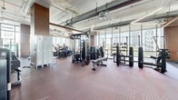 Located at 125 NE 32nd St in Miami's vibrant Upper Eastside neighborhood, this generously sized fitness center delivers a comprehensive, professional-grade workout environment across a single open floor. The space is defined by its dramatic floor-to-ceiling windows that wrap around multiple sides of the room, flooding the interior with abundant natural light and offering sweeping views of the surrounding Miami skyline and cityscape.

The layout is thoughtfully organized into distinct functional zones. A dedicated strength training area features power racks, barbell stations, and a full dumbbell rack, complemented by rubber flooring and lifting platforms designed for heavy use. An extensive selection of selectorized cable machines and resistance equipment anchors the central zone, while a cardio section lines the window-facing wall with a row of treadmills and other aerobic machines positioned to take full advantage of the outdoor views.

Additional amenities include full-length wall mirrors that enhance the sense of openness, built-in storage lockers, and an exposed industrial-style ceiling with polished ductwork that adds an urban, modern aesthetic. Warm wood accent columns contrast with the cool gray tones of the walls, creating a balanced and motivating atmosphere. The facility is well-equipped for a wide range of fitness needs, from functional training to traditional strength and conditioning.