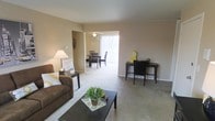 This single-floor residence offers an open and connected layout, with the living area flowing seamlessly into the dining space. The living room is finished with wall-to-wall carpeting in a neutral tone, providing a warm and comfortable foundation for the space. Soft beige walls contribute to an understated, welcoming ambiance throughout.

The living area accommodates a functional arrangement, with room for seating, a coffee table, and a dedicated workspace along one wall, making it versatile for both relaxing and working from home. Decorative wall accents add a touch of character without overwhelming the clean aesthetic.

Adjacent to the living room, the dining area benefits from abundant natural light streaming in through large vertical blinds that line a wide sliding glass door or window, creating a bright and airy atmosphere. A ceiling fan is mounted above the dining space, adding both practical airflow and a subtle design element.

The consistent carpeting and neutral color palette unify both areas, creating a cohesive and harmonious interior. The open sightlines between the living and dining zones enhance the sense of spaciousness throughout this single-level home.