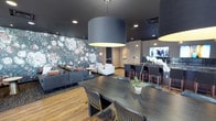 This single-floor communal space is thoughtfully designed to balance style and functionality across multiple distinct zones. At the heart of the room, a striking oversized drum pendant light anchors the dining area, where a dark wood table surrounded by mixed seating provides an ideal setting for gatherings or casual meals. The space flows seamlessly into a comfortable lounge area, featuring a plush sofa and coordinating armchairs arranged around a low side table. A dramatic floral wallpaper mural serves as a bold focal point, adding depth and a touch of nature-inspired elegance to the seating zone. Warm wood-look flooring runs throughout, grounding the design and complementing the dark, moody ceiling treatment. To one side, a bar counter with high stools and pendant lighting creates a social hub, complete with a mounted television for entertainment. Recessed lighting supplements the pendant fixtures, ensuring the entire space is well-illuminated. The overall ambiance is contemporary and refined, with a carefully curated palette of deep charcoals, warm neutrals, and botanical accents that work together to create an atmosphere that is both welcoming and visually dynamic.