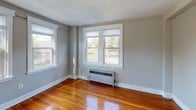 This single-floor unit offers a well-organized layout comprising multiple living spaces, a kitchen, and a bathroom, all connected by a cohesive design aesthetic rooted in classic residential character. Rich, honey-toned hardwood floors run throughout the main living and bedroom areas, reflecting natural light and adding warmth to each room.

The living spaces are bright and open, featuring neutral gray walls, white trim, and multiple double-hung windows framed with clean white molding. Radiator heating units are present in the windowed rooms, consistent with traditional building construction. One room features a distinctive arched doorway opening, adding an architectural detail that lends the space additional charm.

The galley-style kitchen is efficiently laid out with warm oak cabinetry lining both walls, granite-look laminate countertops, a stainless undermount sink, white tile backsplash, and white gas range. The kitchen is finished with durable vinyl plank flooring, providing a practical transition from the hardwood in the adjoining rooms.

The bathroom includes classic black tile accent borders along the walls, a white vanity with sink, a separate shower enclosure, and stone-look tile flooring. White-framed doors with brass hardware add a traditional finish. The dollhouse view confirms a compact but functional floor plan with clear room delineation and good flow throughout.