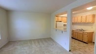 This 2-floor space features a practical and functional layout with a clean, neutral aesthetic. The main living area is finished with plush wall-to-wall carpeting in a light beige tone, creating a soft and comfortable atmosphere. Smooth white walls and a flat ceiling contribute to a bright, airy feel throughout the room.

A defining architectural feature is the pass-through window connecting the living area to the kitchen, which enhances the sense of openness between the two spaces. The kitchen is equipped with warm-toned wood cabinetry, a white refrigerator, and a gas or electric range. The pass-through also incorporates a raised countertop ledge that functions as a breakfast bar, supported by a lower cabinet section. The kitchen flooring transitions to a tile or vinyl surface, distinguishing it from the carpeted living area.

Additional cabinetry is visible in a secondary section of the kitchen, suggesting ample storage and workspace. The overall color palette is consistent and understated, with beige and white tones dominating throughout, lending the space a clean and move-in-ready appearance across both floors.