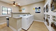 This shared laundry facility in Glendale, Arizona offers a well-organized and efficiently designed space for residents. The room features light beige walls and durable wood-look flooring that lend a clean, neutral aesthetic throughout. Multiple top-load washing machines are arranged in a row along one side, while stacked front-load washer and dryer units line the back wall, maximizing the use of available space and providing ample laundry capacity. A built-in folding counter runs along the perimeter, offering convenient surface space for sorting and folding laundry. An additional wall-mounted shelf provides extra storage for laundry essentials. The facility is equipped with a modern digital payment kiosk, allowing residents to pay via a mobile app—with signage noting a complimentary first wash promotion. Recessed lighting and a ceiling fan ensure the space remains bright and comfortable. A large window along one wall allows natural light to fill the room and offers a view of the surrounding outdoor greenery. Safety is addressed with a mounted fire extinguisher. Overall, the laundry room is clean, practical, and thoughtfully appointed to meet everyday residential needs.