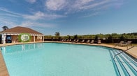 Located at 56 Bent Branch Loop Rd in Clayton, North Carolina, this single-story community clubhouse and amenity area offers residents a well-appointed outdoor retreat. At the heart of the space is a generously sized swimming pool with crystal-clear blue water, surrounded by an expansive concrete pool deck lined with lounge chairs — ideal for relaxing under the North Carolina sun. The pool area is enclosed by a black iron fence, with a backdrop of lush wooded tree lines that provide a sense of privacy and natural beauty.

The clubhouse building itself is designed with classic Southern charm, featuring stone and brick exterior accents, neutral lap siding, and a pitched roof with a cupola. A covered porch with vaulted wood-paneled ceilings and white columnar supports anchored by stone bases extends from the building, creating a shaded outdoor living area. This covered patio is furnished with cushioned seating arrangements and ceiling fans for year-round comfort.

A standout feature of the covered patio is the full-height stone fireplace with a rustic wood mantel, serving as an impressive architectural focal point perfect for cooler evenings. French doors with transom windows connect the patio to the interior clubhouse, allowing natural light to flow through. The overall ambiance is welcoming, refined, and community-oriented.