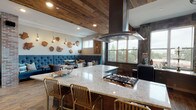 This clubhouse showcases a thoughtfully curated design that merges rustic warmth with industrial sophistication. The kitchen is a standout centerpiece, featuring a generous granite island with a built-in gas cooktop, a sleek overhead range hood, and gold-accented bar stools that invite casual seating. Stainless steel appliances line the perimeter beneath dark, textured wall finishes, complemented by wood-look tile flooring and abundant natural light streaming through large windows that frame outdoor views of the surrounding community and greenery.

The adjacent lounge area offers plush, tufted blue velvet banquette seating alongside mixed dining chairs, creating a comfortable and inviting gathering space. A striking wall display of natural wood slice art adds organic character, while reclaimed wood ceiling planks overhead lend rustic texture and warmth throughout.

A separate mezzanine-level lounge features exposed brick columns, glass-top high tables with industrial-style stools, and dramatic starburst pendant lighting in gold. Colorful artwork anchors the space, adding a vibrant focal point. Black-framed glass doors open directly to an outdoor terrace, seamlessly connecting interior and exterior spaces. Together, these areas create a dynamic, well-appointed clubhouse suited for both casual socializing and larger gatherings.
