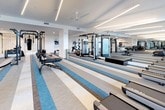 This expansive fitness center is designed with both functionality and aesthetics in mind, offering a comprehensive workout environment across a single, open floor. The space is flooded with natural light through large floor-to-ceiling windows along the perimeter, creating an energizing atmosphere throughout the day. Sleek linear LED lighting runs along the white ceilings, complemented by ceiling fans that ensure comfortable air circulation during workouts.

The flooring features a striking blue, grey, and beige striped carpet in the training zones, transitioning to warm hardwood near the lounge-style bar counter — a stylish centerpiece outfitted with modern pendant lights and metal bar stools, ideal for post-workout refreshment or socializing.

The cardio section lines the windowed wall with a generous row of treadmills, ellipticals, and stair climbers, all equipped with personal display screens. The strength and functional training area is equally well-appointed, featuring a multi-station cable machine, a power rack, a punching bag, battle ropes, and a flat bench. Floor-to-ceiling mirrors amplify the sense of space and assist with form during training. The overall design is clean, modern, and purposefully organized for a premium fitness experience.