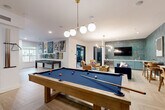 Located at 138 Greison Trail in Newnan, Georgia, this thoughtfully designed single-floor amenity space offers a well-rounded recreational experience. The open layout is anchored by a herringbone-patterned wood-look tile floor that flows seamlessly throughout, lending warmth and visual interest to the entire area.

At the heart of the space sits a full-size billiards table beneath a modern globe pendant chandelier, complemented by a shuffleboard table positioned near large windows that draw in natural light. A comfortable lounge area features a plush navy velvet sofa, tan armchairs, and a vibrant area rug, all set against richly textured teal botanical wallpaper that adds depth and personality.

A dedicated wet bar with sage green cabinetry and a mosaic tile backsplash provides a stylish serving station, while a wall-mounted flat-screen TV makes the lounge ideal for entertaining. A café-style dining nook showcases tufted banquette seating in cognac leather, white tabletops, and bold pendant lighting—ideal for casual gatherings. Curated artwork is displayed throughout, adding a gallery-like ambiance.

An adjacent glass-walled fitness room is visible from the main lounge, housing cardio equipment for residents' convenience. The overall design balances vibrant color with modern sophistication, creating an inviting and versatile community gathering destination.