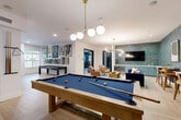 Located at 138 Greison Trail in Newnan, Georgia, this thoughtfully designed single-floor amenity space offers a well-rounded recreational experience. The open layout is anchored by a herringbone-patterned wood-look tile floor that flows seamlessly throughout, lending warmth and visual interest to the entire area.

At the heart of the space sits a full-size billiards table beneath a modern globe pendant chandelier, complemented by a shuffleboard table positioned near large windows that draw in natural light. A comfortable lounge area features a plush navy velvet sofa, tan armchairs, and a vibrant area rug, all set against richly textured teal botanical wallpaper that adds depth and personality.

A dedicated wet bar with sage green cabinetry and a mosaic tile backsplash provides a stylish serving station, while a wall-mounted flat-screen TV makes the lounge ideal for entertaining. A café-style dining nook showcases tufted banquette seating in cognac leather, white tabletops, and bold pendant lighting—ideal for casual gatherings. Curated artwork is displayed throughout, adding a gallery-like ambiance.

An adjacent glass-walled fitness room is visible from the main lounge, housing cardio equipment for residents' convenience. The overall design balances vibrant color with modern sophistication, creating an inviting and versatile community gathering destination.