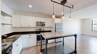 Located on the 8th floor at 1214 Griswold Street in the heart of downtown Detroit, this thoughtfully designed unit offers a well-appointed single-floor layout featuring an open-concept kitchen and living area, one bathroom, and stylish finishes throughout. The living area is characterized by eye-catching herringbone-patterned hardwood floors and large, black-framed windows that flood the space with natural light and frame expansive views of the Detroit skyline. The neutral, warm-toned walls complement the bright, airy atmosphere throughout. The kitchen is a standout feature, boasting white cabinetry, dark granite countertops, stainless steel appliances including a refrigerator, range, microwave, and dishwasher, and open metal shelving. A substantial stainless steel island on casters anchors the space, while industrial-style pendant lights hanging from a pipe fixture add a modern, urban character. The bathroom is clean and contemporary, featuring marble-look tile flooring, a dark vanity with a light countertop, and a glass-enclosed walk-in shower with white subway tile. The overall design balances modern industrial elements with warm, inviting finishes, making this downtown Detroit unit a distinctive and functional urban living space.
