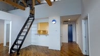 This loft-style space showcases a striking blend of industrial character and modern design. The main living area commands attention with dramatically high ceilings adorned with exposed wooden beams, lending a warm, rustic contrast to the crisp white walls. Rich, wide-plank hardwood floors run throughout, adding texture and a sense of historic charm.

A sleek industrial steel staircase rises along one wall, leading to an elevated loft area above — ideal for additional living or sleeping space. Beneath the loft, a compact kitchen is neatly integrated into the wall, featuring a pass-through counter with seating, light wood cabinetry, and a warm golden tile backsplash that adds a pop of color.

A bold circular blue wall sconce serves as a decorative focal point in the main living area, reinforcing the space's eclectic personality. A corridor leads to the entrance, highlighted by a distinctive deep blue door that makes a strong architectural statement. Recessed lighting and exposed ductwork on the ceiling further emphasize the space's industrial-modern aesthetic.

Overall, this space offers an open, airy atmosphere with thoughtful design details that balance raw, industrial elements with clean, contemporary finishes — creating a dynamic and characterful environment.