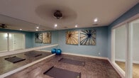 Located at 3501 Farmington Dr in Greensboro, NC, this single-floor yoga and fitness studio is purpose-built for wellness and movement. The main practice room is generously proportioned, offering ample space to accommodate multiple participants simultaneously, with several yoga mats laid out across the durable, stone-toned flooring. Soft blue walls create a calm and focused atmosphere, while full-wall mirrors along two sides of the room visually expand the space and allow practitioners to monitor their form during sessions. Recessed lighting throughout provides even, adjustable illumination, complemented by a ceiling fan that aids in air circulation during active classes. Decorative starburst-style wall art adds an artistic touch that complements the studio's tranquil aesthetic. Stability balls and additional equipment are available within the room. A large glass panel opening leads to an adjacent entry and restroom area, which features light hardwood-style flooring, a restroom with accessibility features, and a fire safety station. The entry area benefits from natural light through an exterior-facing door, connecting the studio to the outdoors. Overall, the space is thoughtfully designed to support a focused, serene environment suitable for yoga, stretching, meditation, or light fitness activities.
