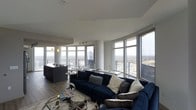 This elegant high-rise residence offers a seamlessly integrated open-concept living space defined by clean lines, contemporary finishes, and an abundance of natural light. Floor-to-ceiling windows wrap around the main living area, framing breathtaking panoramic views of the city skyline and surrounding landscape — a true centerpiece of the home.

The living area is anchored by a generously sized navy sectional sofa, complemented by layered textiles and a glass-and-metal coffee table that adds a refined, modern touch. The rich wood-look flooring runs throughout, providing warmth against the crisp white ceilings and neutral walls.

The kitchen flows directly into the living space in true open-plan fashion, featuring a large dark-toned island that serves as both a prep area and casual gathering spot. Track lighting overhead ensures the kitchen is well-lit and functional, while pendant lights add a subtle decorative accent.

Large sliding glass doors lead to an outdoor balcony, extending the living space and offering an elevated vantage point over the cityscape. The overall ambiance is sleek, cosmopolitan, and light-filled — ideal for those who appreciate modern urban living with a dramatic backdrop.