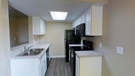 This single-floor space is designed with functionality and comfort in mind. The kitchen serves as a standout feature, laid out in a classic galley configuration that maximizes efficiency. White shaker-style cabinetry lines both sides of the kitchen, offering generous storage space above and below the countertops. The light-toned countertops provide a clean contrast against the sleek black appliances, which include a refrigerator, electric range, built-in microwave, and dishwasher. A double-basin stainless steel sink is positioned along one wall, complemented by additional counter space for meal preparation. A large skylight overhead floods the kitchen with natural and ambient light, brightening the entire space. The warm wood-look flooring runs throughout, adding a cohesive and inviting feel. The neutral beige wall tones create a versatile backdrop that complements the overall aesthetic. The layout is thoughtfully designed to make the most of the available space, ensuring ease of movement and practicality in everyday use. Overall, the space presents a clean, move-in-ready environment with a balanced blend of modern fixtures and timeless design elements.