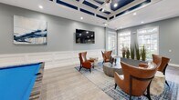 Located at 1301 Ivy Meadow Dr in Charlotte, NC, this single-floor clubhouse amenity space combines sophisticated design with a welcoming, social atmosphere. The main lounge area is anchored by a striking coffered ceiling with navy blue accents and recessed lighting, creating an elevated sense of volume and refinement. Wainscoting along the walls, light hardwood-style flooring, and a soft gray color palette lend the space a polished, cohesive aesthetic. Comfortable leather wingback chairs arranged around sculptural concrete side tables invite relaxed gatherings, while a wall-mounted flat-screen television adds a functional entertainment element. A statement billiards table with a bold blue felt surface serves as a focal point for recreation and socializing. Large French doors and expansive windows flood the interior with natural light while offering views of the surrounding outdoor environment. The entry area features striking large-format stone tile flooring alongside structural columns that define separate zones without closing off the open layout. A dedicated kitchenette area includes dark cabinetry with gold hardware, white countertops, and a wine refrigerator, making it well-suited for hosting events. Together, these elements create a dynamic, upscale communal space designed for both leisure and entertainment.
