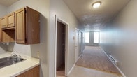 This single-floor residential unit offers a straightforward and functional layout designed for comfortable everyday living. A central hallway with neutral carpet runs the length of the space, connecting key areas and providing an open, accessible flow throughout. The hallway is lined with doors leading to separate rooms, with a built-in closet featuring wire rack shelving providing ample organized storage. Adjacent to the closet, wood-toned cabinetry with a light countertop surface adds practical utility, ideal for storage or workspace use. The textured ceiling runs consistently throughout, contributing to the overall character of the interior. At the end of the hallway, a well-lit room benefits from a large window that draws in natural light, brightening the space considerably. A baseboard heater beneath the window ensures comfortable climate control year-round. The neutral gray wall tones and beige carpeting throughout create a clean, understated palette that is easy to personalize. Overall, this unit presents a practical and well-organized single-level layout with functional storage solutions and consistent finishes, making it a solid and livable space.