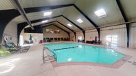 Located at 175 La Fontenay Dr in Louisville, Kentucky, this single-level indoor pool facility is a standout recreational amenity. The centerpiece of the space is a generously sized lap pool, prominently branded with the "La Fontenay" name on its floor, filled with clear turquoise water. The structure's most striking architectural feature is its dramatic vaulted ceiling, supported by bold dark structural beams that rise to a steep peak. Multiple skylights are integrated into the ceiling, flooding the interior with natural light and creating an open, airy atmosphere. The walls are finished in light vertical board paneling, lending the space a clean and classic aesthetic. Surrounding the pool, a wide concrete deck provides ample space for lounging, furnished with black metal chaise lounges, dining tables, and chairs arranged throughout. Sliding windows and glass doors along the perimeter offer views of the surrounding green grounds, enhancing the connection to the outdoors. Ceiling fans assist with ventilation and climate comfort. A raised secondary area with additional seating is visible at one end of the pool, adding dimension to the layout. The overall ambiance is bright, functional, and welcoming, making it well-suited for both recreational swimming and relaxed social gatherings.