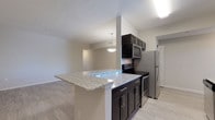This single-floor residence offers a thoughtfully designed layout with modern finishes throughout. The kitchen is a standout feature, configured as a galley-style space with dark espresso cabinetry, light granite countertops, and a full suite of stainless steel appliances including a range, microwave, and refrigerator. A peninsula with matching granite extends into the adjacent living area, providing a natural division between the kitchen and the open living and dining space. Light wood-style flooring runs throughout the main living areas, complementing the neutral gray-toned walls and creating a bright, cohesive aesthetic. Pendant lighting adds a decorative touch above both the peninsula and the dining area. The bedroom features soft wall-to-wall carpeting and a closet with built-in rod storage. The bathroom includes a soaking tub with dark tile surround, a vanity with dark cabinetry and a light countertop, and a large mirror. A dedicated laundry area houses a stacked washer and dryer unit with a window providing natural light. Overall, the space presents a clean, contemporary feel with consistent finishes.