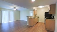 This single-floor space is designed around an open-concept layout that seamlessly connects the kitchen and living area. The kitchen features a peninsula with granite-style countertops, an undermount sink, light maple cabinetry, and stainless steel appliances, including a built-in dishwasher and refrigerator. Recessed and flush-mount ceiling lighting provides consistent illumination throughout the kitchen zone. The living area extends naturally from the kitchen, highlighted by rich, dark hardwood-style flooring that runs throughout the space and a ceiling fan that adds both function and a casual aesthetic. Soft, neutral green-toned walls complement the warm wood tones, creating a cohesive and welcoming atmosphere. A large window in the living area allows generous natural light to fill the room, enhancing its open feel. White baseboards and clean trim details add a polished finish throughout. The single-floor layout ensures easy flow between areas, making the space practical and functional for everyday living.