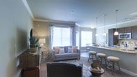 This single-floor residence is designed with an open-concept layout that seamlessly connects the living area and kitchen, creating a cohesive and functional living environment. The living room is highlighted by large windows dressed with light, patterned curtains that allow generous natural light to fill the space, enhancing the neutral, warm-toned walls accented by classic crown molding. The room comfortably accommodates a well-appointed seating arrangement, including a plush sofa with decorative accent pillows and additional lounge seating, all centered on a stylish area rug. A media console and decorative greenery add personality to the space. Adjacent to the living area, the kitchen features a granite or stone-topped breakfast bar with bar stool seating, making it ideal for casual dining and entertaining. Pendant lighting hangs above the bar, providing warm, focused illumination. A dining area is visible in the background, nestled near additional windows that further brighten the interior. The overall ambiance is modern yet comfortable, with a cohesive color palette of soft grays, taupes, and warm neutrals throughout, offering a welcoming and polished aesthetic suited for everyday living.