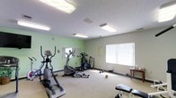 This single-floor fitness room provides a dedicated space for cardiovascular exercise and general workouts. The room is finished with light green walls and neutral-toned carpet flooring, creating a clean and functional environment. Overhead fluorescent lighting ensures the space is well-illuminated throughout, supplemented by natural light from a window fitted with vertical blinds along one wall.

The room is equipped with a selection of cardio machines, including an elliptical trainer, a stair climber, and a stationary bike, arranged to allow comfortable movement between equipment. A wall-mounted mirror near one corner enhances the sense of space and provides a visual reference during workouts. Additional accessories, including dumbbells and a weight bench, are also present, offering basic strength training options.

A small side table and informational postings on the walls contribute to the practical layout of the room. Ventilation is provided through ceiling vents, supporting a comfortable workout environment. Overall, this fitness room is a straightforward, well-maintained space designed to meet everyday exercise needs.