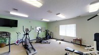 This single-floor fitness room provides a dedicated space for cardiovascular exercise and general workouts. The room is finished with light green walls and neutral-toned carpet flooring, creating a clean and functional environment. Overhead fluorescent lighting ensures the space is well-illuminated throughout, supplemented by natural light from a window fitted with vertical blinds along one wall.

The room is equipped with a selection of cardio machines, including an elliptical trainer, a stair climber, and a stationary bike, arranged to allow comfortable movement between equipment. A wall-mounted mirror near one corner enhances the sense of space and provides a visual reference during workouts. Additional accessories, including dumbbells and a weight bench, are also present, offering basic strength training options.

A small side table and informational postings on the walls contribute to the practical layout of the room. Ventilation is provided through ceiling vents, supporting a comfortable workout environment. Overall, this fitness room is a straightforward, well-maintained space designed to meet everyday exercise needs.