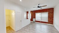 Located at 1057 S Western Ave in Los Angeles, CA, this single-floor unit offers a compelling blend of classic architectural character and modern updates. The main living area immediately draws attention with its striking exposed red brick accent wall, which adds warmth and texture against the clean white walls and ceiling. Light wood-look flooring runs throughout, creating a bright and cohesive feel underfoot. A ceiling fan provides both function and style overhead, while recessed lighting ensures the space is well-lit. Windows fitted with white blinds invite natural light while maintaining privacy. A wall-mounted heater serves the living area efficiently. The kitchen is thoughtfully appointed with white shaker-style cabinetry, brushed nickel hardware, gray countertops, a deep single-basin sink, and a gas range, offering a clean and functional cooking space. The bathroom features a full soaking tub and shower combination, with elegant marble-patterned wall surrounds in white and gray tones, complemented by a small window for ventilation and natural light. The layout flows intuitively between rooms, with additional doors leading to closet or storage space. Overall, the unit presents a refined yet approachable aesthetic that balances historic charm with contemporary convenience in the heart of Los Angeles.