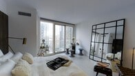 This stylish high-rise residence offers a refined living experience defined by clean lines, a crisp neutral palette, and an abundance of natural light. The bedroom is a standout feature, showcasing expansive floor-to-ceiling windows that frame breathtaking panoramic views of the surrounding urban skyline, with neighboring towers and open sky creating a dramatic backdrop. Light-toned flooring runs throughout, enhancing the sense of openness and airiness in the space. The room is thoughtfully appointed with contemporary furnishings, including sculptural side stools and a striking black industrial-framed mirror that adds depth and visual interest to the white walls. A balcony or window access point allows for an extended connection to the outdoors and the cityscape beyond. The minimalist aesthetic is cohesive and intentional, with carefully curated décor — including fresh floral arrangements and layered textiles — that adds warmth without compromising the space's modern sensibility. The smooth white ceilings and uncluttered layout contribute to an overall ambiance that is both calming and sophisticated, making this a serene urban retreat elevated above the city below.