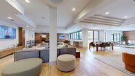 This expansive single-floor amenity space offers a thoughtfully designed open-concept layout that seamlessly integrates multiple functional zones across a generous footprint. Light hardwood flooring flows throughout the main gathering areas, complemented by warm wood-paneled accent walls that add texture and visual warmth against a crisp white palette. Recessed lighting and coffered ceiling details provide architectural interest overhead, while structural columns punctuate the open floor plan without disrupting the spacious feel.

The lounge area features plush sectional seating, upholstered ottomans, and wall-mounted televisions, creating a comfortable setting for relaxation and socializing. Rustic wood stump side tables add organic contrast to the contemporary furnishings. A dedicated game area includes a full-size pool table, while a separate dining zone provides seating for groups around a dining table. Additional co-working or casual seating areas are positioned near floor-to-ceiling windows, which frame outdoor views and flood the space with natural light.

A secondary area features tiled flooring with tufted seating, banquette-style booths upholstered in teal, and curated artwork. Private rooms with glass-panel doors offer quieter, flexible spaces. Throughout, the design balances a modern aesthetic with welcoming, residential warmth.