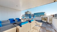 Located at 1801 Chestnut Pl in Denver, Colorado, this rooftop terrace offers a well-appointed outdoor amenity space with a modern, urban aesthetic. The deck is finished with a patterned wood tile surface that adds warmth and texture underfoot, creating a cohesive look throughout the space. A partially covered lounge area provides shade and comfort, furnished with wicker seating accented by vibrant blue cushions, arranged around a central coffee table — ideal for relaxed gatherings or casual conversation. Moving into the open-air section of the terrace, an additional seating arrangement accompanies a bar-height dining table and chairs, also outfitted in blue, providing a dedicated space for outdoor dining. A built-in fireplace serves as a focal point in the open deck area, adding both ambiance and functionality for cooler Denver evenings. The terrace is bordered by low perimeter walls that open up to panoramic views of the surrounding Denver skyline, including nearby residential and commercial buildings. The bright, airy atmosphere is enhanced by abundant natural sunlight and clear blue skies, making this rooftop an inviting destination for both daytime relaxation and evening entertaining in the heart of the city.