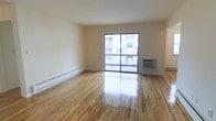 This single-floor unit presents a clean, open living area centered around polished hardwood floors that reflect natural light throughout the space. The main living area is bright and uncluttered, with neutral cream-toned walls that complement the warm wood flooring. A full-width sliding glass door serves as a focal point, allowing generous amounts of natural light to fill the room while providing direct access to a balcony with views of the surrounding building exterior. Practical climate control is well-addressed, with a wall-mounted air conditioning unit positioned near the sliding door and baseboard heating running along the perimeter walls — ensuring year-round comfort. The layout is straightforward and versatile, offering flexibility for various furniture arrangements. The overall ambiance is light, clean, and functional, making it a practical and livable space suitable for a range of occupants.