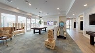 Located at 6600 Rolling Meadows Dr in Sparks, Nevada, this single-level community amenity space is designed to deliver an engaging and social atmosphere for residents. The layout flows across distinct yet connected zones, blending entertainment, relaxation, and community interaction seamlessly.

The main game area is generously sized and well-lit, featuring wood-look plank flooring and patterned carpet tiles that define different activity zones. A full-size pool table serves as a central focal point, flanked by a mounted flat-screen TV and wall art. Nearby, a skee-ball arcade machine, oversized Jenga, and giant Connect Four add a playful, activity-rich character to the space.

Adjacent to the game area, a shuffleboard table sits beneath a trio of large, wall-mounted screens — ideal for watching sports or programming while playing. A bar-height counter with backless stools provides a casual seating and viewing area, while a pass-through connects to what appears to be a mailroom or service corridor.

A separate lounge area offers a more relaxed ambiance, centered around a floor-to-ceiling metal-clad fireplace wall with a modern inset firebox. Comfortable wingback and accent chairs surround the hearth, and rustic barn-style double doors add a warm, character-rich architectural detail. Natural light filters in through glass doors leading to an outdoor area, further enhancing the open, welcoming feel of the space.
