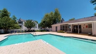 This outdoor pool area offers a welcoming and well-maintained recreational retreat. The centerpiece is a generously sized swimming pool with crystal-clear turquoise water, clearly marked with depth indicators and no-diving zones for safety. The pool is surrounded by a broad, light-toned paver deck that provides ample space for lounging chairs and umbrellas, encouraging outdoor relaxation under open skies. A white perimeter fence encloses the area, adding both security and a clean, classic aesthetic. Mature trees line the boundary of the space, offering natural shade and a lush green backdrop that enhances the overall ambiance. Adjacent to the pool is a single-story clubhouse-style building with white lap siding, large multi-pane windows, and classic blue double doors — the structure provides a shaded covered porch area supported by painted columns, creating a comfortable spot to rest out of direct sunlight. Colorful plantings and decorative container pots add welcoming touches of color throughout the patio surroundings. The space is equipped with emergency safety features, including a wall-mounted emergency phone. The combination of open sky, natural greenery, and thoughtfully designed hardscaping makes this pool area a bright and inviting outdoor amenity.
