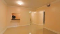 This single-floor space offers a clean, functional layout designed for comfortable everyday living. The main living area is finished with highly polished flooring that reflects light throughout the room, contributing to an open and airy atmosphere. Warm, neutral-toned walls painted in a soft peach hue lend the space a welcoming feel, while crisp white crown molding adds a refined architectural detail along the ceiling perimeter. A textured ceiling with flush-mount lighting provides consistent illumination across the living area. A notable feature is the kitchen pass-through window, fitted with a dark countertop ledge, which connects the kitchen to the main living space and enhances the sense of openness while maintaining a practical separation between areas. White baseboards run throughout, complementing the overall clean aesthetic. A hallway visible from the main room leads further into the space, suggesting additional rooms beyond. The layout flows naturally, making efficient use of the available footprint. Overall, the space presents a tidy, well-maintained interior with classic finishing touches that create a bright and inviting environment suitable for a variety of uses.