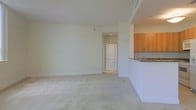 This single-floor residence offers a clean, well-lit interior defined by neutral tones and a thoughtful open layout. The main living area is generously proportioned, featuring plush wall-to-wall carpeting and smooth white walls that reflect natural and ambient light to create a bright, airy atmosphere. The space feels open and unobstructed, lending itself well to a variety of furniture arrangements.

Adjoining the living area is a kitchen that is partially separated by a raised breakfast bar counter finished with a stone or granite surface, adding a touch of texture and elegance. The kitchen features warm honey-toned wood cabinetry that provides ample storage, complementing the lighter palette of the rest of the space. A sink is visible beneath the upper cabinets, and the overall kitchen design balances functionality with a welcoming aesthetic.

A doorway from the main living area leads further into the unit, suggesting additional rooms beyond. The interior door features a classic arched panel design, adding a subtle architectural detail that enhances the overall character of the space. Crisp white baseboards and trim run throughout, tying the rooms together with a cohesive, polished finish. The space presents a well-maintained, move-in-ready environment with a calm, neutral ambiance suitable for a range of interior styles.