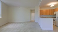 This single-floor residence offers a clean, well-lit interior defined by neutral tones and a thoughtful open layout. The main living area is generously proportioned, featuring plush wall-to-wall carpeting and smooth white walls that reflect natural and ambient light to create a bright, airy atmosphere. The space feels open and unobstructed, lending itself well to a variety of furniture arrangements.

Adjoining the living area is a kitchen that is partially separated by a raised breakfast bar counter finished with a stone or granite surface, adding a touch of texture and elegance. The kitchen features warm honey-toned wood cabinetry that provides ample storage, complementing the lighter palette of the rest of the space. A sink is visible beneath the upper cabinets, and the overall kitchen design balances functionality with a welcoming aesthetic.

A doorway from the main living area leads further into the unit, suggesting additional rooms beyond. The interior door features a classic arched panel design, adding a subtle architectural detail that enhances the overall character of the space. Crisp white baseboards and trim run throughout, tying the rooms together with a cohesive, polished finish. The space presents a well-maintained, move-in-ready environment with a calm, neutral ambiance suitable for a range of interior styles.