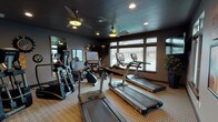 Located at 13101 Shetland Lane in Spokane, Washington, this dedicated fitness center offers a well-equipped, single-floor workout facility designed for both cardio and strength training. The space is finished with a neutral-toned textured ceiling and warm gray walls, creating a focused yet inviting atmosphere. Recessed lighting and dual ceiling fans provide ample illumination and air circulation throughout.

The cardio section is generously stocked with multiple TRUE-brand treadmills and elliptical trainers, as well as upright stationary bikes, all arranged to allow comfortable use. A large floor-to-ceiling mirror enhances the sense of space and supports proper form during workouts. Windows along the perimeter allow natural light to filter in, adding brightness to the room.

On the strength side, a multi-function cable machine and a dedicated weight bench provide options for resistance training. A built-in water fountain station offers convenient hydration access, and a wall-mounted rack keeps accessories organized. Decorative touches — including framed artwork, sunburst wall mirrors, and starburst accent pieces — lend the space a polished, community-oriented aesthetic. The carpeted flooring runs throughout, providing cushioning underfoot for a comfortable workout environment.