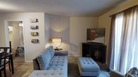 This single-floor space offers a functional and inviting layout centered around a well-appointed living area. The living room serves as the heart of the space, featuring a plush tufted sectional sofa in a soft blue-grey upholstery, complemented by a matching tufted ottoman that adds both comfort and versatility. A sleek corner media console provides a practical anchor for the entertainment setup, while a stylish arc table lamp casts warm ambient lighting throughout the room. The walls are finished in neutral tones, creating a calm and adaptable backdrop, and are accented with decorative wall signage that adds a touch of personality and character. The textured ceiling adds subtle architectural interest overhead. Carpeted flooring runs throughout, contributing to the overall warmth and comfort of the space. The corner layout of the living area makes efficient use of the available square footage, offering a cozy yet open feel. Overall, the space presents a straightforward, livable environment with thoughtful furnishings and a casual, approachable aesthetic well-suited for everyday use.