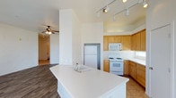 This single-floor space is designed with an open-concept layout that promotes a sense of airiness and connectivity throughout. The kitchen serves as the heart of the home, anchored by a large central island with a built-in sink and expansive white countertops — ideal for meal preparation and casual gatherings. Warm honey-toned wood cabinetry lines the walls, complemented by white appliances including a gas range, built-in microwave, and refrigerator. Track lighting runs along the ceiling, providing focused illumination across the kitchen workspace. The flooring transitions from tile in the kitchen to rich wood-look planks in the adjoining living area, creating a natural visual distinction between the two spaces. Soaring ceilings enhance the open feel of the home, while a ceiling fan in the living area ensures comfortable airflow year-round. The architectural design emphasizes clean lines and neutral tones throughout, creating a bright and welcoming ambiance. The generous layout and thoughtful flow between kitchen and living areas make this space both functional and inviting for everyday living.