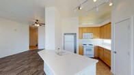 This single-floor space is designed with an open-concept layout that promotes a sense of airiness and connectivity throughout. The kitchen serves as the heart of the home, anchored by a large central island with a built-in sink and expansive white countertops — ideal for meal preparation and casual gatherings. Warm honey-toned wood cabinetry lines the walls, complemented by white appliances including a gas range, built-in microwave, and refrigerator. Track lighting runs along the ceiling, providing focused illumination across the kitchen workspace. The flooring transitions from tile in the kitchen to rich wood-look planks in the adjoining living area, creating a natural visual distinction between the two spaces. Soaring ceilings enhance the open feel of the home, while a ceiling fan in the living area ensures comfortable airflow year-round. The architectural design emphasizes clean lines and neutral tones throughout, creating a bright and welcoming ambiance. The generous layout and thoughtful flow between kitchen and living areas make this space both functional and inviting for everyday living.