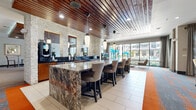 Located at 1616 W Dallas St in Houston, TX, this sophisticated clubhouse and amenity space delivers a resort-inspired experience for residents. The interior design is defined by a striking slatted wood ceiling that runs throughout, creating warmth and visual continuity across each area. Stacked stone accent walls provide a natural, textural contrast against the contemporary finishes, while bold, patterned carpeting in shades of gray and orange grounds the social spaces with energy and style.

The welcoming entry lobby features decorative bamboo accents, a resident services desk, and a glass floor feature that adds an architectural focal point. The lounge area offers generous seating with a sectional sofa, upholstered chairs, and a flat-screen television mounted against a stacked stone wall — ideal for relaxed gathering.

The game room is a standout amenity, outfitted with a full-size billiards table, a shuffleboard table, and bistro-style high-top seating. Floor-to-ceiling windows flood the space with natural light and offer views of the outdoor pool area, maintaining a vibrant connection to the exterior.

A granite-topped bar with pendant lighting and cushioned bar stool seating provides a stylish social hub, complemented by large windows that frame the surrounding Houston skyline. Throughout, the space balances refined materials with a welcoming, community-centered atmosphere.