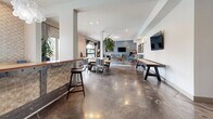 Located at 400 Buford Hwy in Suwanee, GA, this single-floor amenity space offers a generously sized, open-concept layout designed for both relaxation and recreation. Polished concrete floors run throughout, lending an industrial-modern aesthetic that ties the entire space together cohesively.

The living room area features a striking floor-to-ceiling matte black accent wall, comfortable seating arrangements, large windows that flood the space with natural light, and views to the outdoors. A warm, layered lighting scheme complements the curated mix of contemporary furnishings.

The game room is a standout highlight, equipped with a billiards table, a ping pong table set atop a decorative area rug, and a shuffleboard table. Industrial-style garage doors open to the exterior, seamlessly blending indoor and outdoor living. Exposed brick-style tile accent walls add texture and warmth, while tall indoor plants contribute a vibrant, organic element throughout.

A built-in banquette dining area with a navy upholstered bench, striped accent chairs, and elegant pendant lighting provides a sophisticated space for communal dining or gatherings. A bar counter with seating offers an additional casual gathering spot. Wall-mounted televisions and thoughtful décor complete this well-appointed, multifunctional community space.