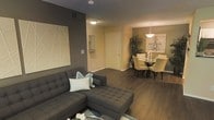 This well-appointed single-floor residence showcases a thoughtfully designed open-concept layout that seamlessly connects the living and dining areas. Rich, dark hardwood flooring flows throughout, anchoring the space with warmth and sophistication.

The living area is defined by a large, tufted charcoal sectional sofa accented with light blue and white throw pillows, complemented by a striking geometric wall art piece mounted against a bold dark accent wall. The contrast between the deep gray tones and the softer neutral walls creates a modern, layered aesthetic.

Flowing naturally from the living space, the dining area is illuminated by an elegant chandelier pendant light that casts a warm ambiance over a round glass-top dining table surrounded by cream upholstered chairs. A large decorative plant adds a touch of organic texture to the corner, enhancing the room's inviting atmosphere.

A well-placed entryway with a white door connects the interior to the outside, featuring a small mirror and hook area — a practical and stylish touch. The overall design palette of warm neutrals, deep charcoals, and rich wood tones gives this residence a contemporary yet comfortable feel, making it an ideal space for both everyday living and entertaining.