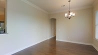 This single-story space showcases a thoughtfully designed interior that balances classic elegance with functional layout. The room featured in the image is finished with rich, dark hardwood flooring that adds warmth and depth to the space. Crown molding lines the ceiling perimeter, lending a refined, traditional character to the room.

A decorative chandelier with a bronze-toned finish and frosted glass shades serves as a focal point, providing both ambient lighting and a touch of sophistication. The walls are painted in a neutral, warm beige tone that complements the flooring and creates a bright, welcoming atmosphere.

A distinctive arched doorway leads through to an adjoining area, adding architectural interest and a sense of continuity between spaces. This arched transition is framed with clean white trim, consistent with the baseboards and crown molding throughout. The ceiling features a visible HVAC vent, indicating a central air system integrated into the home's infrastructure.

Overall, the space presents a clean, move-in-ready interior with classic detailing that suits a variety of décor styles, from traditional to transitional.
